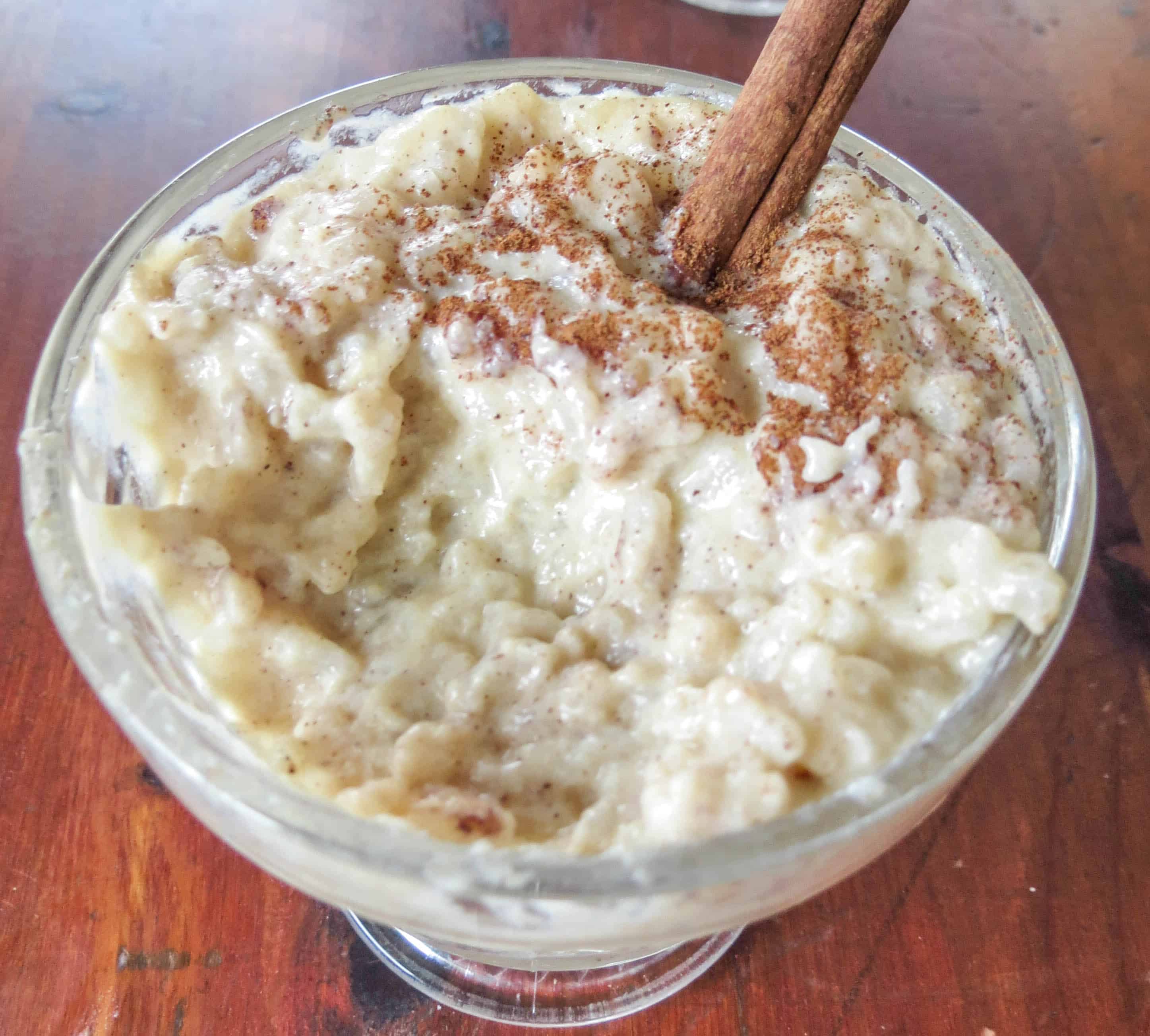 Homemade creamy rice pudding in a clear glass cup with cinnamon sprinkled on top and a cinnamon stick stuck in. There is a bite mixing to reveal texture.