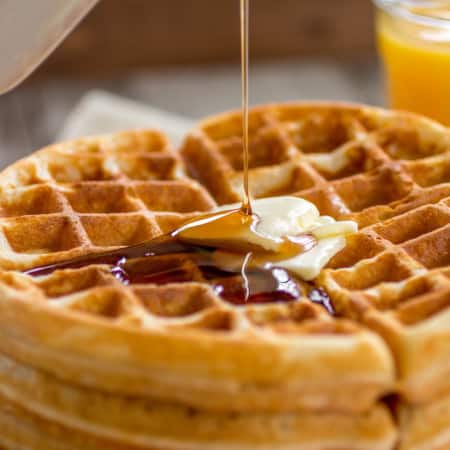 Waffles stacked on a plate with a pat of butter on top getting syrup poured on top.