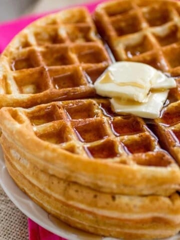 Waffles stacked on a plate with syrup and butter.