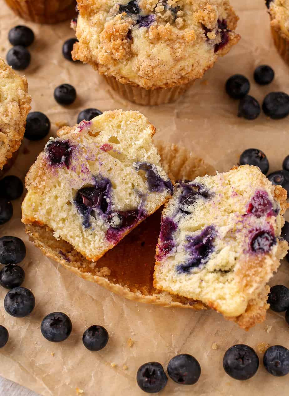 Blueberry Muffin cut in half on brown parchment paper with blueberries scattered around.