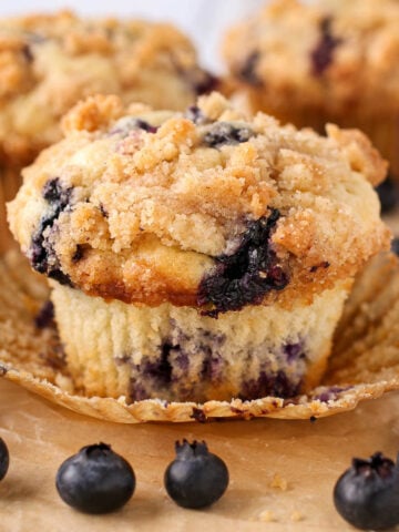Blueberry crumb muffin with streusel topping with wrapper unwrapped and blueberries scattered around it.