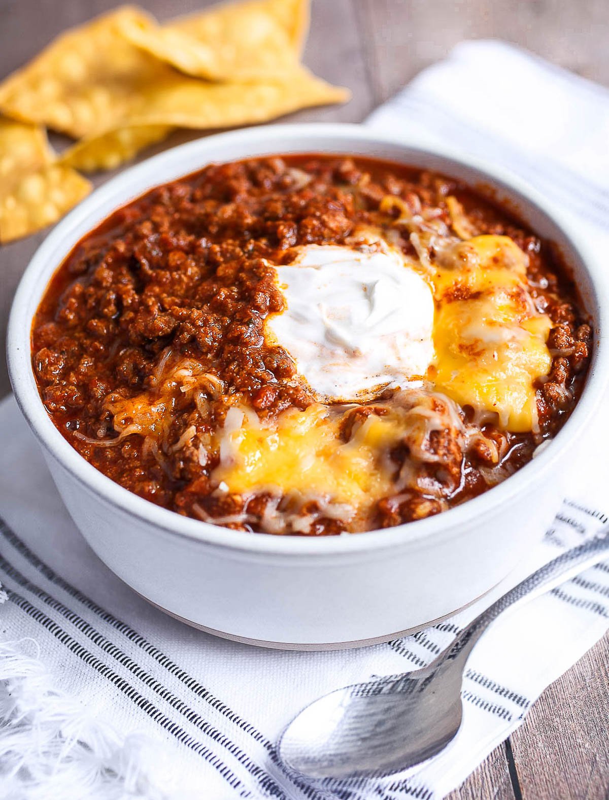 Classic bowl of chili recipe with a dollop of sour cream and melted cheese on top with chips in the background.